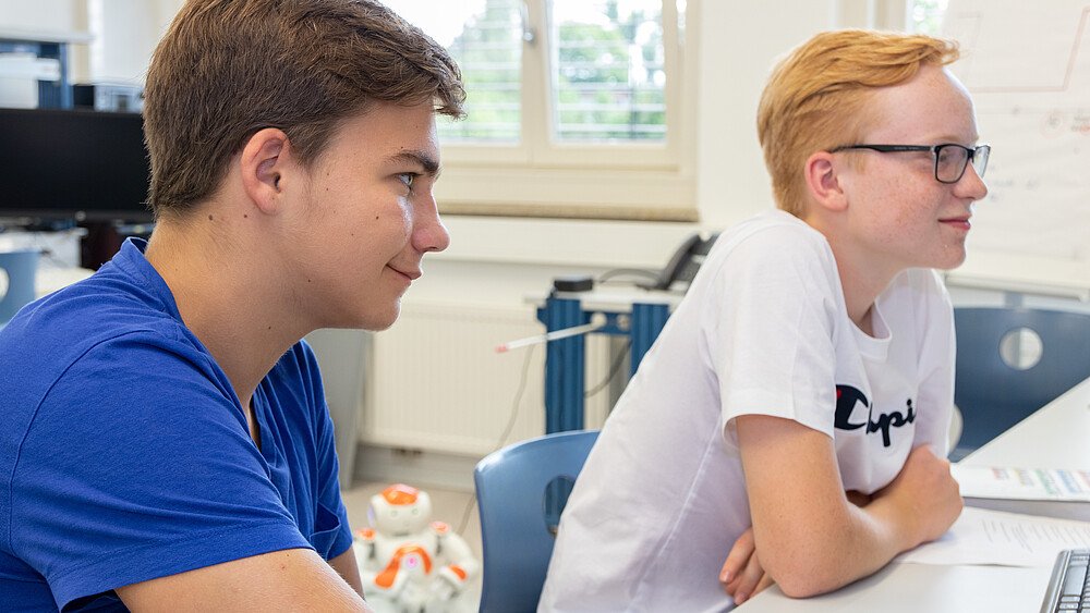 Zwei Schüler sitzen am Tisch in einem Labor und blicken in Richtung Rednerpult.