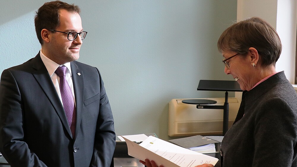 A middle-aged man with a purple tie and black suit, wearing a white shirt underneath and glasses, is presented with a certificate by a woman. A pastel blue-green wall can be seen in the background.