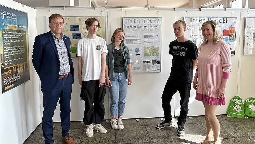The dean, two students, one student and the vice-rector stand in front of white back walls with posters on the topic of sustainability pinned to them and smile into the camera.