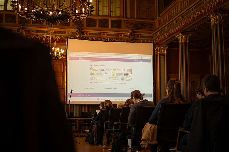 Guests sit on rows of chairs in the citizens' hall of Zittau town hall. A beamer projection on a screen shines in the background.