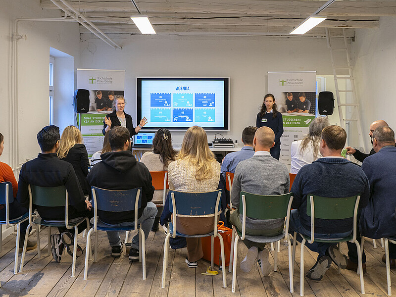 Workshop participants sit on rows of chairs and listen to two moderators.