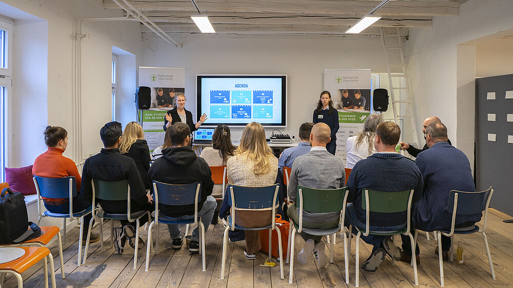 Workshop participants sit on rows of chairs and listen to two moderators.