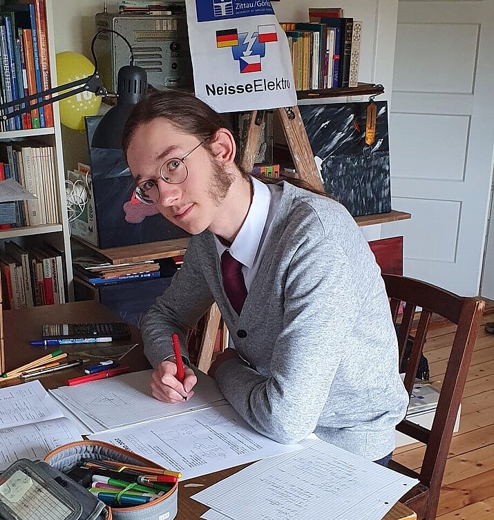 A student sits at his desk, pen in hand. Behind him hangs a Neisse-Elektro poster.