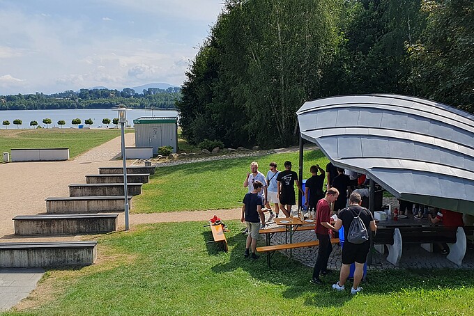 A collection of adult people standing by a lake under a shell-shaped roof