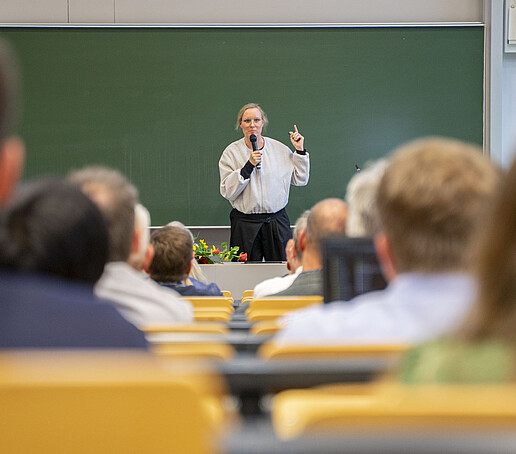 A woman speaks into a microphone in front of a green board in the lecture hall.