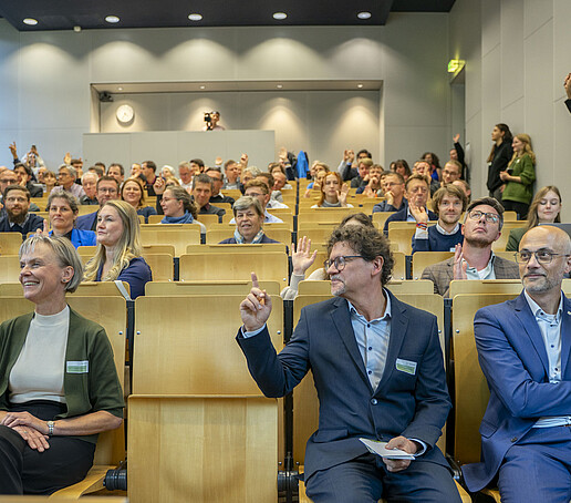Rows of seats in the lecture hall are filled with audience members. Many are holding their hands in the air.