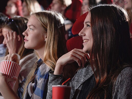 Several young people sit in the movie theater, popcorn in hand, smiling as they watch the screen.
