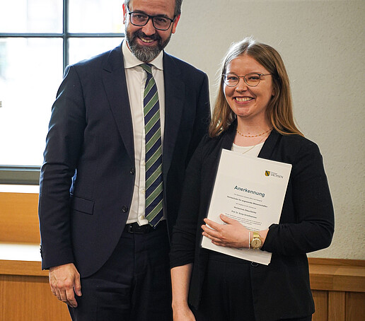 A man and a woman stand next to each other, both smiling at the camera. The woman is holding a certificate with the logo of the Free State of Saxony.