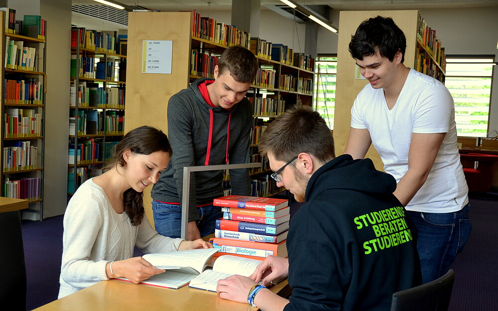 Four students are in the library. One SbS student points to a text lying on the table in front of him. The others listen with interest. A student is leafing through a notebook that is open in front of her.