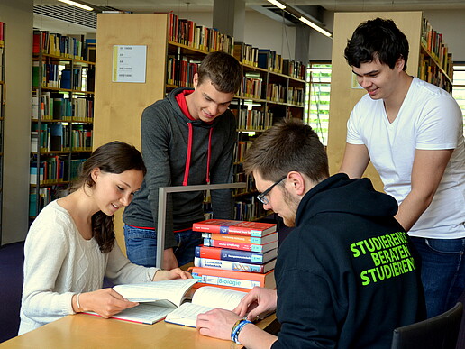 Four students are in the library. One SbS student points to a text lying on the table in front of him. The others listen with interest. A student is leafing through a notebook that is open in front of her.