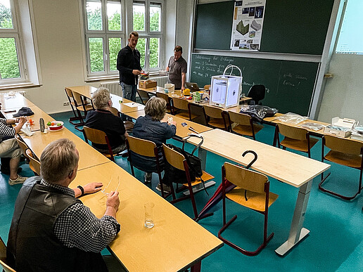 Citizens sit at desks and look at the blackboard. Two university employees are standing there explaining the principle of a 3D printer.