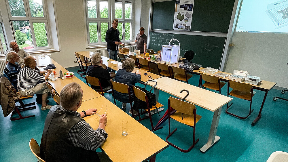 Citizens sit at desks and look at the blackboard. Two university employees are standing there explaining the principle of a 3D printer.