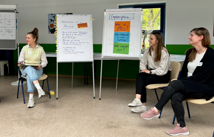 Three female students sit in a semicircle in front of display stands that are labeled and partly covered with notes.