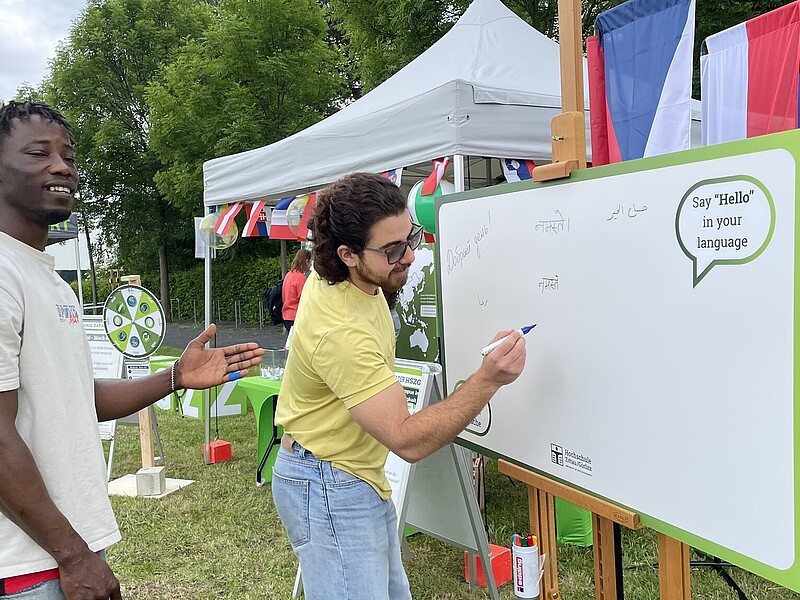 Two international students are standing in front of a blackboard. One of them is writing a greeting on the board with a pen.