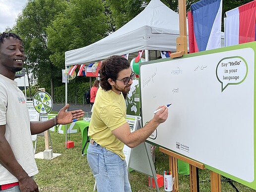 Two international students are standing in front of a blackboard. One of them is writing a greeting on the board with a pen.