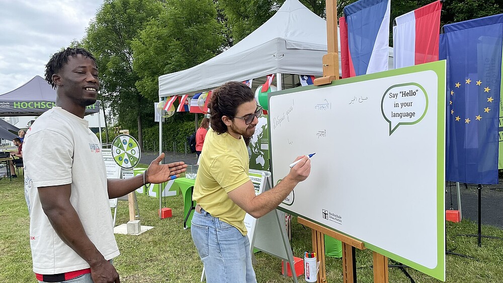 Two international students are standing in front of a blackboard. One of them is writing a greeting on the board with a pen.