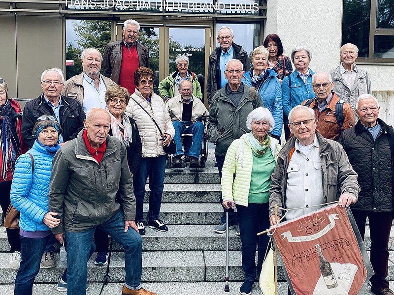 Eine Gruppe ehemaliger Studierender steht auf den Treppenabsätzen vor Haus eins. Ein älterer Herr hält eine Studiumsflagge in der Hand. Alle schauen in die Kamera.
