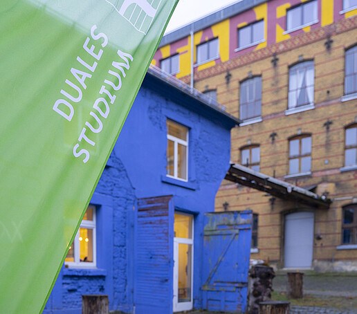 A brick building stands in the background, with a blue house with white shutters to the left. A beach flag with the inscription "Duales Studium" is waving in the foreground.