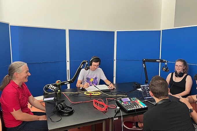 Podcast participants sit at a table surrounded by sound walls. The recording equipment is on the table.