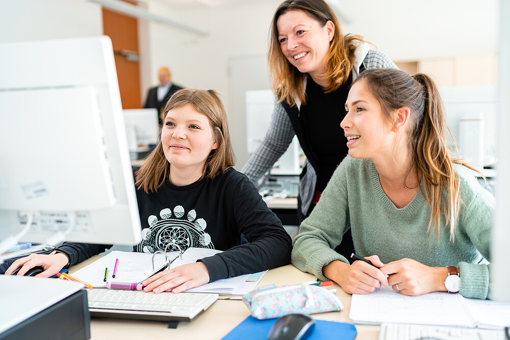 Two students are sitting in front of the computer. The professor stands behind them and explains things.
