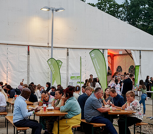 Vor dem Festzelt sitzen die vielen Gäste auf Bänken und feiern.