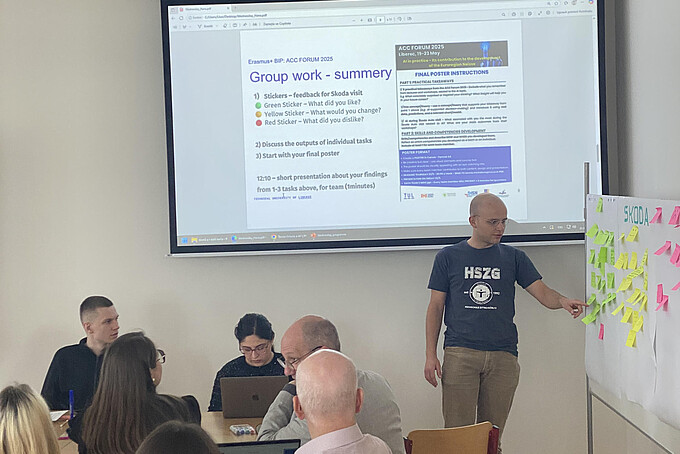 A student explains something to a group on a display with colorful cards. A presentation appears on a screen in the background.