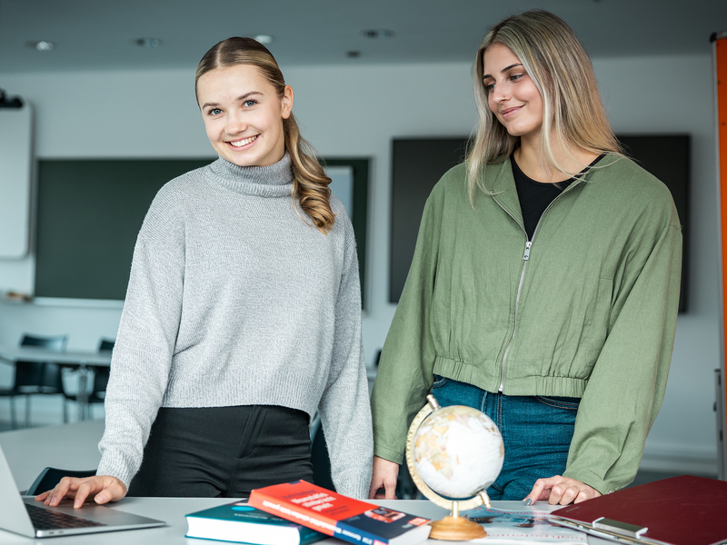 Zwei Studentinnen stehen an einem Tisch mit aufgeklappten Laptop. Vor ihnen liegen Studienmaterialien.