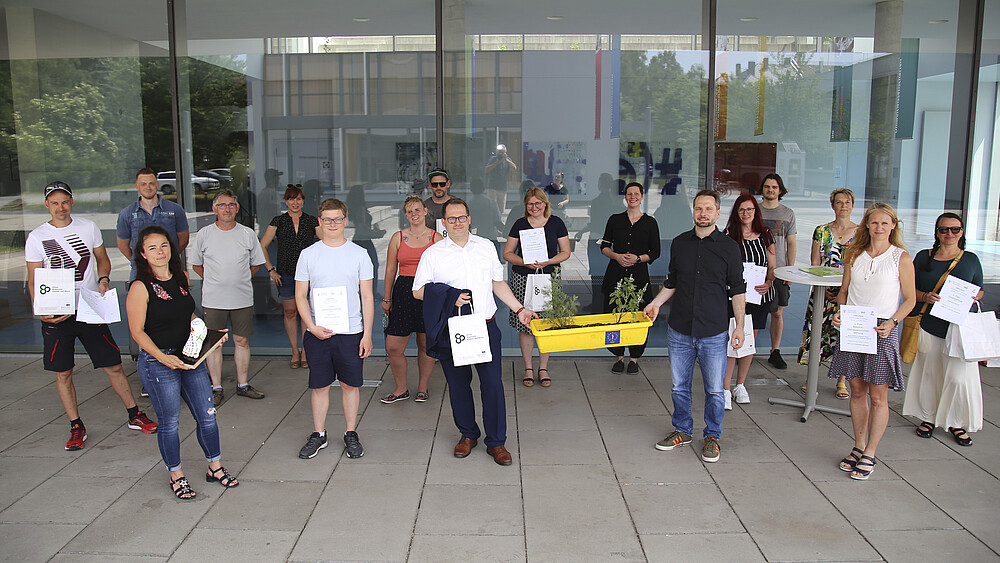 Eine große Gruppe von Schrittzähler-Challenge-Teilnehmenden steht vor Haus 4 und lächelt in die Kamera. Die Ausgezeichneten halten ihre Urkunden in die Kamera.