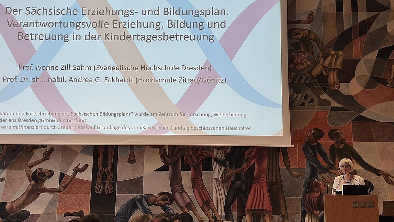 Professorin Eckhardt steht an einem Rednerpult, daneben ist eine Projektionsfläche, auf der eine Präsentation läuft.