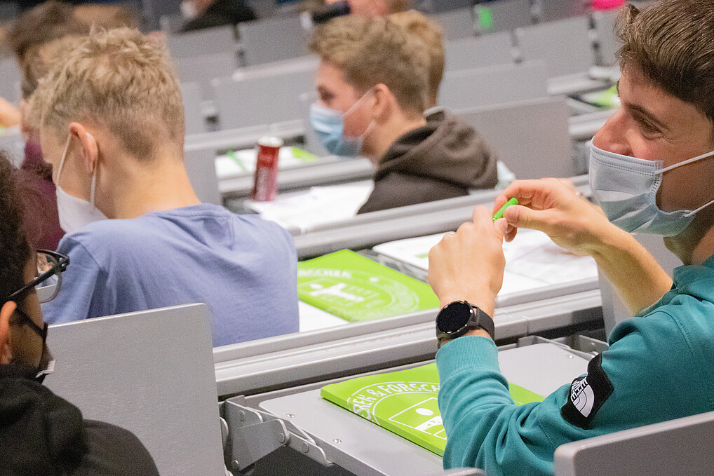 Lachende Studierende sitzen im Hörsaal und tragen Mund-Nasen-Schutzmasken.