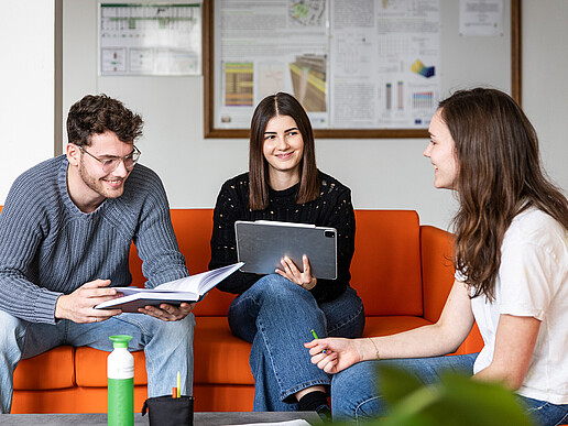 Three students sit on an orange sofa and talk.