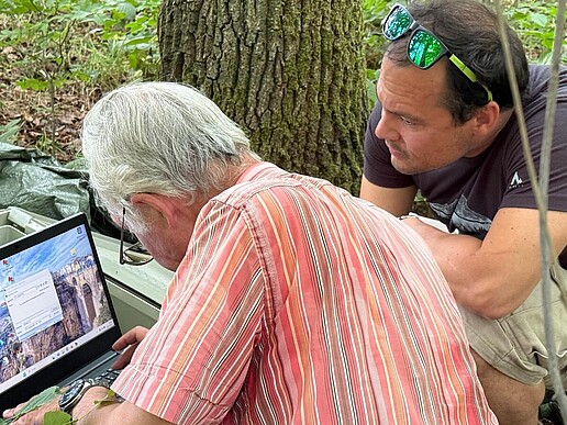 Prof. Hermann Heilmeier (TU Freiberg) and Prof. Oliver Wiche (HSZG) are sitting on the ground in front of an open metal case in a wooded area near Freiberg. Heilmeier is operating a laptop, while Wiche looks attentively over his shoulder. Trees and the forest floor can be seen in the background. The scene shows the readout of a data logger as part of a Europe-wide measurement campaign on soil water balance and drought stress in trees, initiated by a call from the Society for Ecology in May 2025.