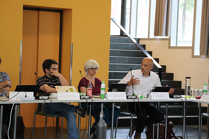 Two deans and a female dean sit at a long table in the auditorium and confer.