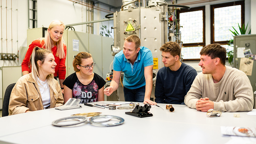 Eine Gruppe Studierender sitzt am Tisch in einem Techniklabor. Ein Lehrender steht dabei und erklärt einen Versuchsaufbau.