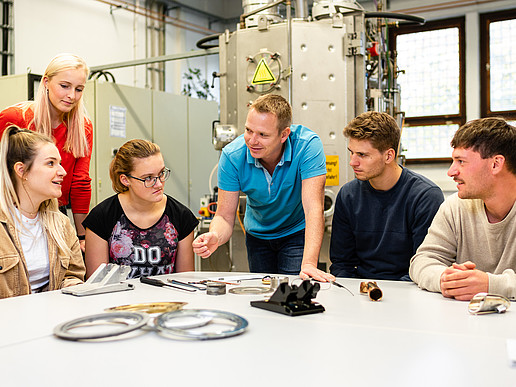 A group of students is sitting at a table in a technology lab. A teacher is standing by and explaining an experimental setup.