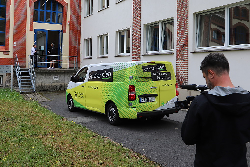 The university bus and a cameraman are standing in front of the Zittau power plant laboratory. The rector and the blogger are standing in front of the entrance talking.