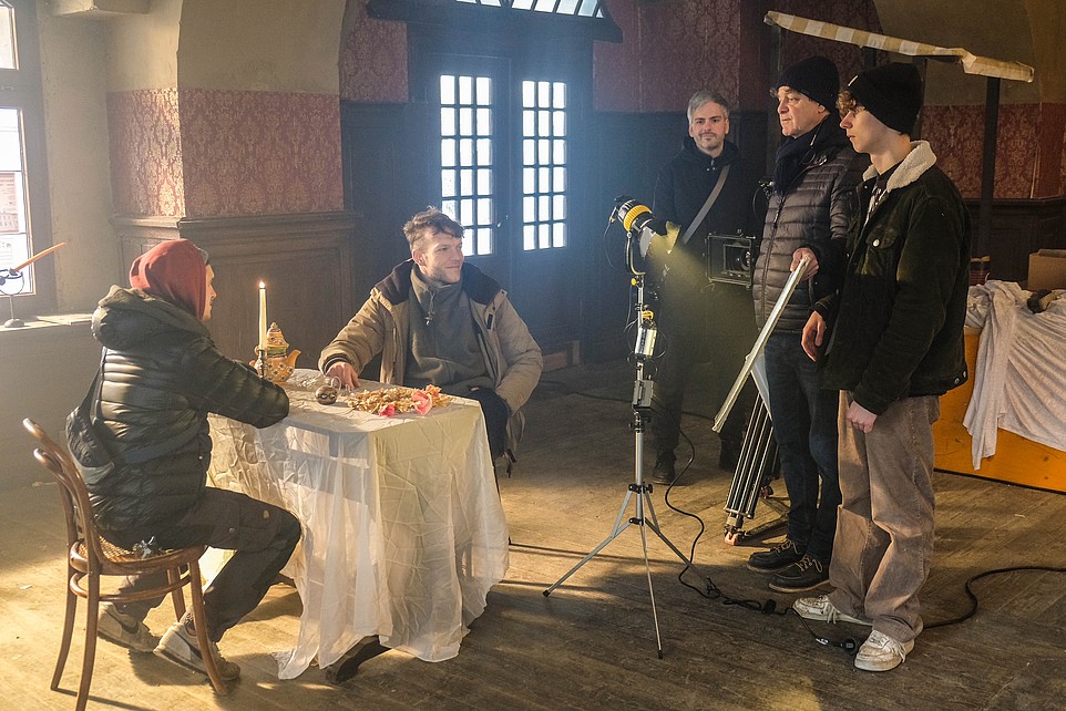 Two course participants sit at a set table in a film set and discuss with the course participants standing next to them.