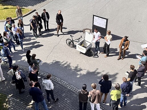 Eine Gruppe Menschen stehen auf einem Hof im Kreis. An der oberen Seite steht ein Fahrrad und ein Blackboard.