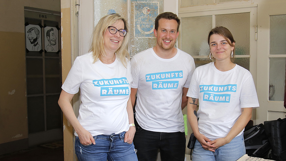 Three people wearing uniform T-shirts with the words "Zukunftsräume" (future spaces) are standing in a fragrance seminar room.