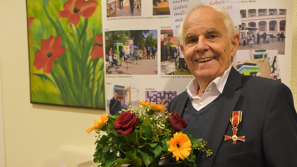 Konrad Kutt wears the Federal Cross of Merit and holds a bouquet of flowers in his hands. He smiles into the camera.