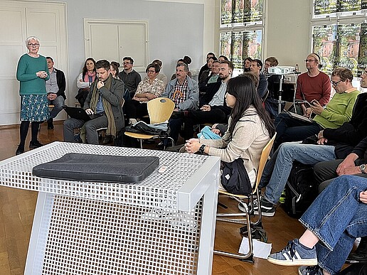 Teilnehmende eines Fachaustauschs sitzen auf Stuhlreihen und hören einer Dozentin zu.