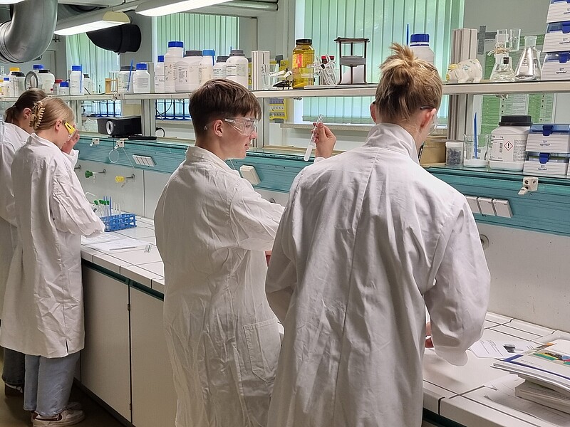 Schüler und Schülerinnen in weißen Kitteln stehen im Labor und experimentieren.
