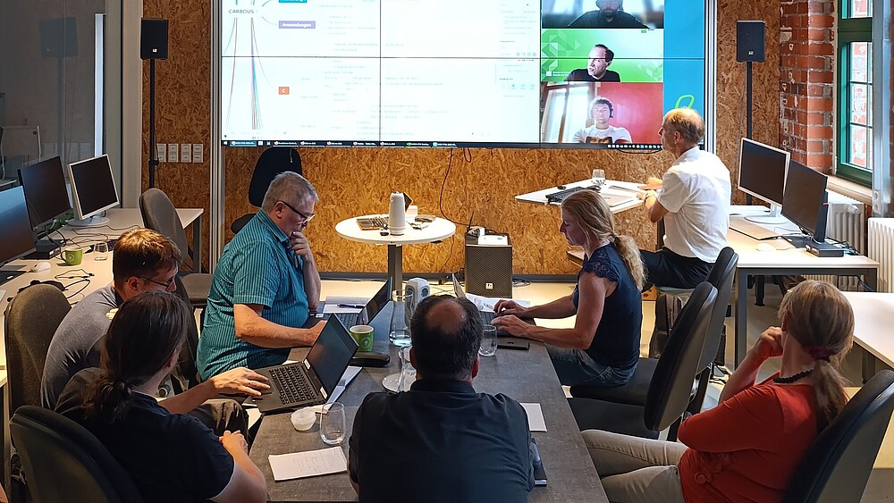 People sit at tables in the Celsiuz, in the background there is a large digital screen on which presentations are displayed and people are connected live via webcam.
