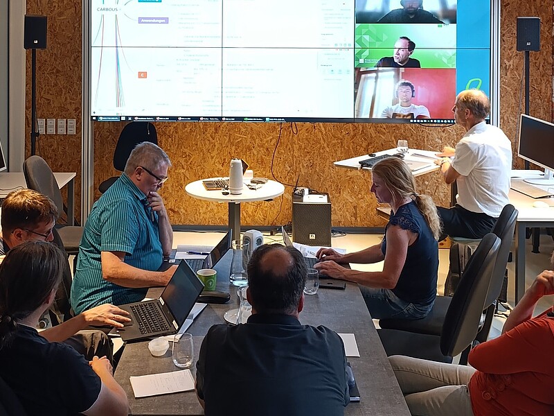 Menschen sitzen an Tischen im Celsiuz, im Hintergrund steht eine große digitale Leinwand, auf der Präsentationen abgebildet und Personen live per Webcam zugeschaltet sind.