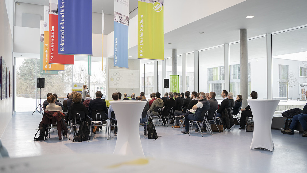 Rows of chairs and high tables are set up in the foyer of House Z IV. The audience seated there listens to a speaker.