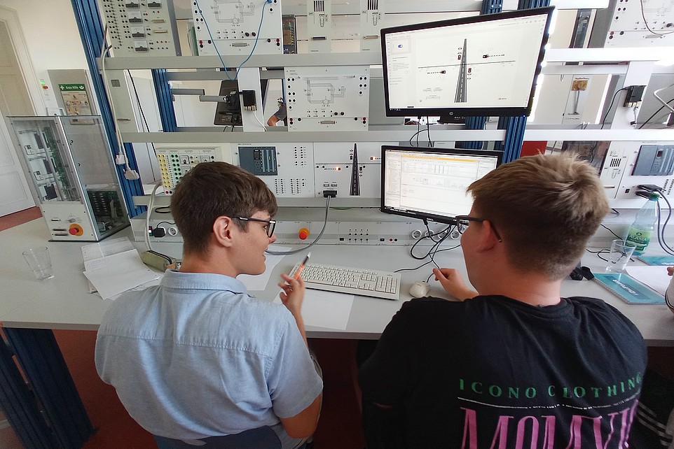 Two young men sit in front of a test stand with several monitors