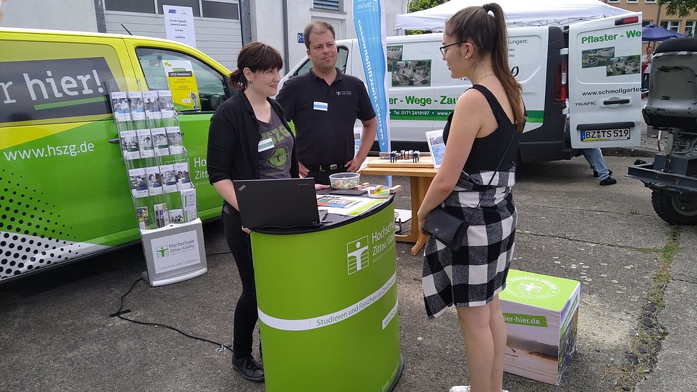 A member of staff and an employee of the HSZG stand at an advice stand and advise a young woman. The university bus can be seen in the background.
