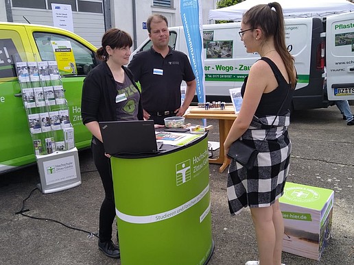 A member of staff and an employee of the HSZG stand at an advice stand and advise a young woman. The university bus can be seen in the background.