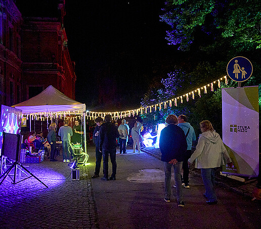 The StuRa stand with roll-up and pavilion and a string of lights, visitors walk past it.