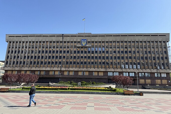 Ein monumentales Verwaltungsgebäude mit mehreren Etagen ist auf dem Vorplatz zu sehen. Viele Fenster sind mit Holzplatten verschlossen, das Gebäude trägt über dem Eingang das ukrainische Wappen. Auf dem Platz davor spazieren vereinzelt Menschen.